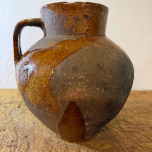 Puede incluir: Una jarra de barro rústica con cuerpo bulboso y una sola asa lateral. La superficie presenta un acabado moteado con parches de esmalte marrón oscuro, ámbar y gris carbón, mostrando una apariencia texturizada y desgastada con signos de uso.