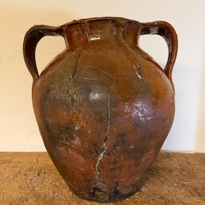 May include: A rustic, bulbous earthenware vessel featuring a deep brown, mottled glaze with visible surface cracks and weathering. The pot has two symmetrical side handles and a wide, flared rim, showcasing a textured, aged appearance.