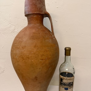 Puede incluir: Una jarra grande y antigua de terracota con asa, junto a una botella de vino. La jarra tiene una base ancha y un cuello estrecho. La botella de vino tiene una tapa dorada y una etiqueta que dice "Chateau de Suduiraut 1929 Bordeaux."