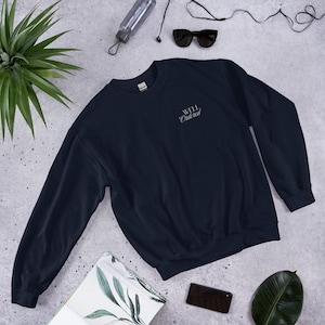 May include: A dark navy blue crewneck sweatshirt featuring the text "WELL ORDERED" embroidered in a white script font on the upper left chest area. The garment has long sleeves, a ribbed collar, and ribbed cuffs, presented in a flat lay view.