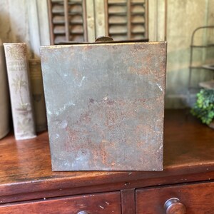 Op de afbeelding: Een vintage metalen doos met een versleten, roestige afwerking. De doos heeft een scharnierend deksel en is rechthoekig. De doos staat op een houten oppervlak.