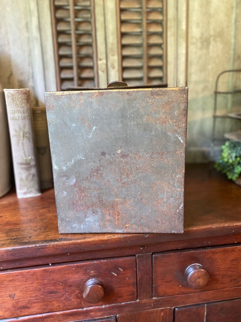 Op de afbeelding: Een vintage metalen doos met een versleten, roestige afwerking. De doos heeft een scharnierend deksel en is rechthoekig. De doos staat op een houten oppervlak.