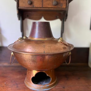 May include: A vintage copper hot pot with a flared base and a chimney-like top. The pot has two handles and a decorative opening on the base. The copper has a warm, aged patina, suggesting its use over time. The pot is designed for cooking and serving.
