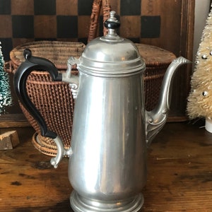 May include: A silver pewter coffee pot with a black handle and a black knob on the lid. The pot is sitting on a wooden surface.
