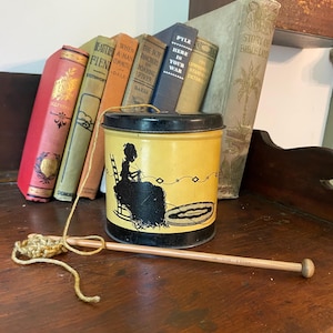 May include: A vintage yellow and black tin can with a silhouette of a woman knitting. The can is sitting on a wooden surface with a wooden knitting needle and yarn next to it. The can has the text "Pyle Here Is Your War" on it.