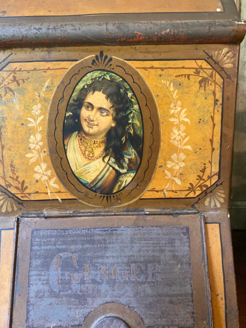 Op de afbeelding: Antiek metalen blik met een geschilderd beeld van een vrouw met lang donker haar, die een ketting en een bloemenkrans draagt. Het blik heeft een gouden achtergrond met bloemmotieven en het woord "GINGER" staat op de voorkant gedrukt.
