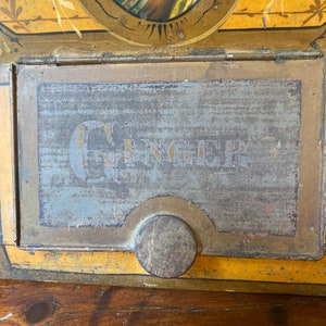 Op de afbeelding: Een vintage metalen blik met een gele en gouden achtergrond. Het blik heeft een geschilderd beeld van een vrouw in een blauwe jurk en een gouden kroon. Het blik heeft een scharnierend deksel met een metalen sluiting en het woord "GINGER" staat op de voorkant gedrukt.