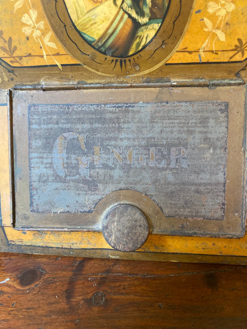 Op de afbeelding: Een vintage metalen blik met een gele en gouden achtergrond. Het blik heeft een geschilderd beeld van een vrouw in een blauwe jurk en een gouden kroon. Het blik heeft een scharnierend deksel met een metalen sluiting en het woord "GINGER" staat op de voorkant gedrukt.