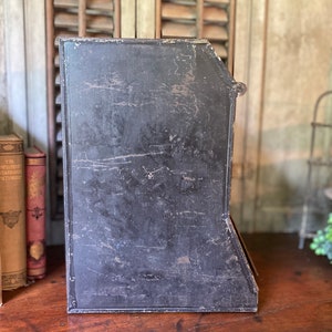 Op de afbeelding: Een zwarte metalen tijdschriftenrek met een versleten, verweerde afwerking. Het rek heeft vijf vakken om tijdschriften of andere items in op te bergen.