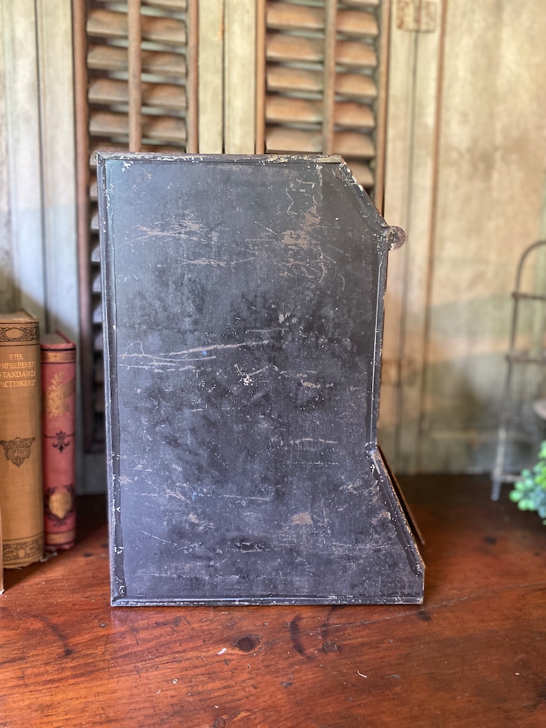 Op de afbeelding: Een zwarte metalen tijdschriftenrek met een versleten, verweerde afwerking. Het rek heeft vijf vakken om tijdschriften of andere items in op te bergen.