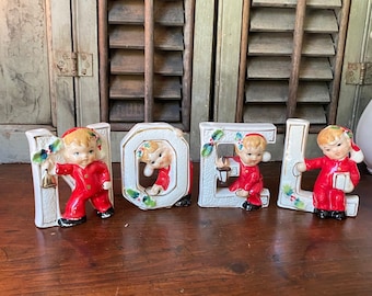 May include: White ceramic letters spelling "NOEL" with a small boy figurine in each letter wearing a red and white suit and a Santa hat. The letters are decorated with green holly leaves and red berries.
