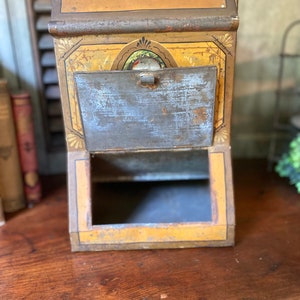 Op de afbeelding: Vintage metalen blik met een geel en gouden geverfd ontwerp. Het blik heeft een scharnierende deur en een kleine opening aan de bovenkant. Het blik staat op een houten oppervlak.