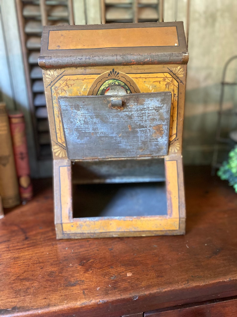 Op de afbeelding: Vintage metalen blik met een geel en gouden geverfd ontwerp. Het blik heeft een scharnierende deur en een kleine opening aan de bovenkant. Het blik staat op een houten oppervlak.