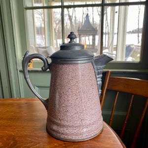 May include: A vintage coffee pot with a textured, reddish-brown body and a dark metal lid and handle. The spout and handle are made of the same dark metal. The pot is sitting on a wooden table.