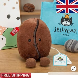 Puede incluir: Un juguete de peluche marrón con forma de grano de café con una cara sonriente y pies pequeños. El juguete tiene una etiqueta que dice "Best Seller". Una bolsa de mano azul con un logotipo de gato negro y las palabras "Jellycat London" en el fondo.
