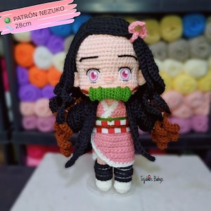 May include: A handmade crochet doll of a character with black hair, pink eyes, and a green mouth guard. The doll is wearing a pink and white kimono with a black jacket. The doll is approximately 11 inches tall and has the text "Patrón Nezuko 28cm" in the upper left corner.