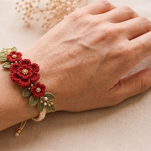 May include: A bracelet featuring three crocheted red flowers with gold bead centers, surrounded by crocheted green leaves and small white pearls. The floral arrangement is attached to a beige braided cord with a sliding knot closure and gold beads.