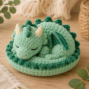 Puede incluir: Un dragón hecho a mano, tejido a ganchillo en tonos verdes, acurrucado en posición de dormir. El dragón tiene los ojos cerrados, cuernos color crema y un cuerpo texturizado. El dragón descansa sobre una base redonda verde.