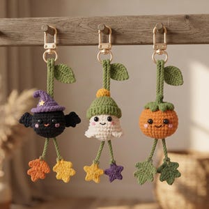 May include: Three crochet keychains hanging from gold-tone clasps. The first is a black bat with a purple witch hat, the second a white ghost in a green beanie, and the third an orange pumpkin. Each features green stems, leaves, and dangling charms.