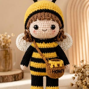 May include: A handmade crochet doll dressed as a bee. The doll has a yellow and black striped outfit, a matching hat with antennae, and small white wings. It carries a brown honey pot. The doll is approximately 8 inches tall.