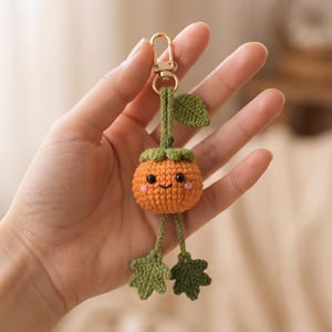 May include: A small, round, orange crocheted pumpkin keychain with a green stem, a single green leaf, and two dangling green legs ending in leaf-shaped feet. The pumpkin features a stitched black smile, two black bead eyes, and pink cheeks.