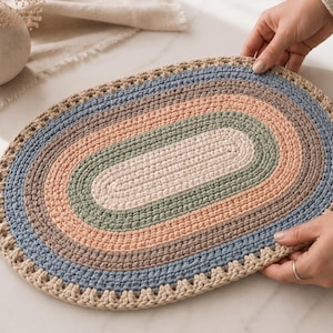 May include: An oval-shaped crochet mat featuring concentric rings of yarn in shades of cream, sage green, dusty pink, brown, and muted blue. The outer edge is finished with a decorative scalloped border in a light cream color.