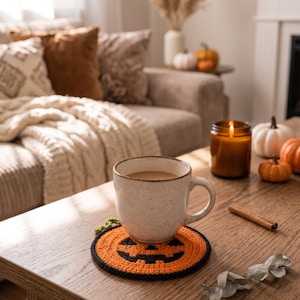 May include: A round, crocheted coaster featuring a jack-o'-lantern face design in orange and black yarn with a small green stem detail. The coaster is placed on a wooden surface and supports a speckled, off-white ceramic mug filled with a light brown beverage.