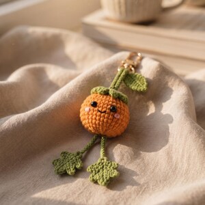 May include: A crocheted orange fruit keychain with a smiling face, black bead eyes, and pink stitched cheeks. The orange has a green stem, a green leaf, and two dangling green legs with leaf-shaped feet, all attached to a gold-toned metal clasp.