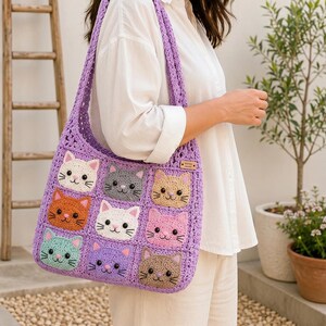 Puede incluir: Bolso tote de crochet hecho a mano en un vibrante tono púrpura. El bolso presenta una cuadrícula de nueve caras de gato en varios colores, incluyendo blanco, naranja y gris. El bolso tiene una correa larga para llevar al hombro o en la mano.