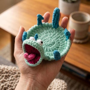 Könnte beinhalten: Ein handgefertigter Häkeluntersetzer in Form eines Cartoon-Monsters. Der Untersetzer ist hauptsächlich hellgrün mit blauen Akzenten, weißen Augen und einem rosa Mund und einer Zunge. Der Untersetzer wird in der Hand einer Person gehalten.