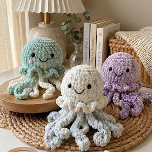 May include: Three handmade crochet jellyfish toys in shades of blue, white, and purple. Each plushie has a smiling face and long, curly tentacles. The toys are displayed on a wooden surface and a woven mat, with books and a lamp in the background.