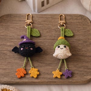 May include: Two handmade crochet keychains featuring a black bat wearing a purple hat and a white ghost wearing a green hat. Both figures have green stems with leaves at the top, gold-tone lobster clasps, and dangling feet shaped like leaves and stars.