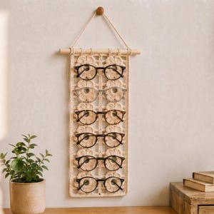 Pode incluir: Um painel de macramé cor de creme com um padrão floral repetitivo e cinco pares de óculos pendurados nas alças tecidas. O organizador está suspenso por uma haste de madeira clara com um cordão a condizer para fixação na parede.