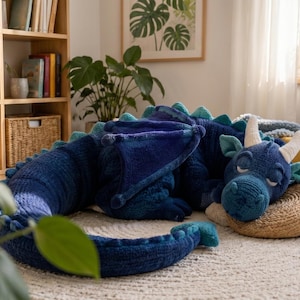 May include: A large, plush, dark blue dragon stuffed animal with a textured, ribbed surface. The dragon features light blue accents along its back spikes, wings, and ears, with two cream-colored horns on its head and closed eyes in a sleeping pose.