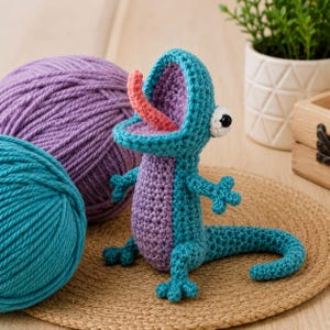Puede incluir: Un lagarto de ganchillo hecho a mano en color turquesa y morado con la boca abierta y una lengua rosa. El lagarto tiene un ojo blanco grande con una pupila negra. Dos ovillos de lana en turquesa y morado están en el fondo.