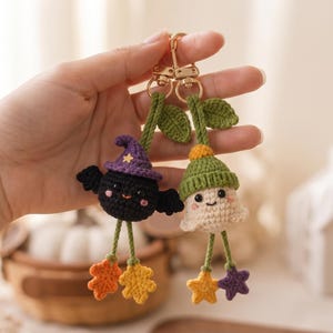May include: Two crochet keychains featuring a black bat wearing a purple witch hat and a white ghost wearing a green hat. Both figures have green leaf accents at the top, dangling legs, and colorful star-shaped feet attached to gold metal clasps.