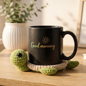 Pode incluir: Uma caneca de cerâmica preta com as palavras "Good morning" e um gráfico de sol, apoiada em um descanso de copo de tartaruga de crochê verde e bege. Um vaso branco com uma planta está no fundo.