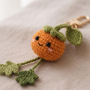 May include: A crocheted orange fruit keychain with a smiling face, black bead eyes, and pink cheeks. It features a green stem with a leaf, two dangling green leaves on thin stems, and a gold-toned metal lobster clasp for attachment.
