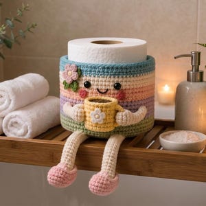 May include: A whimsical, handmade crochet toilet paper holder in pastel colors. The holder has a smiling face, arms, and legs, holding a yellow mug. The holder is decorated with a flower and is sitting on a wooden shelf with rolled white towels.