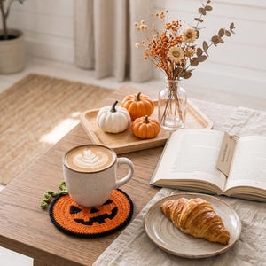 May include: A round, crocheted coaster featuring an orange jack-o'-lantern face with a black border and a small green stem. The coaster is placed under a speckled ceramic mug filled with coffee, next to a croissant on a plate and an open book.
