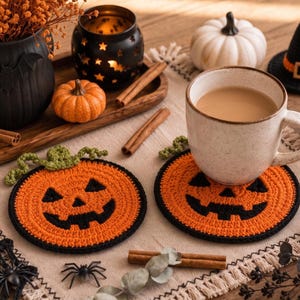 May include: Two round, orange and black crocheted coasters featuring a jack-o'-lantern face design with a green crocheted stem. The coasters are crafted with a textured stitch pattern and a black border, designed to hold a beverage mug.