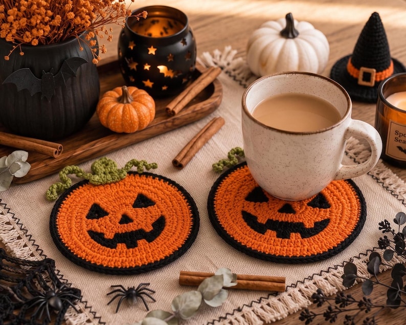 May include: Two round, orange and black crocheted coasters featuring a jack-o'-lantern face design with a green crocheted stem. The coasters are crafted with a textured stitch pattern and a black border, designed to hold a beverage mug.