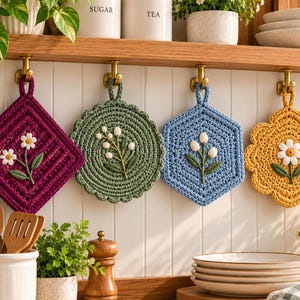 May include: Four crocheted pot holders hang from brass hooks. The set includes a purple square, a green circle, a blue hexagon, and a yellow flower shape. Each piece features a central embroidered floral design with white petals and green leaves.