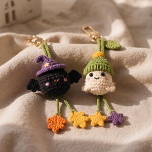 May include: Two crochet keychains featuring a black bat wearing a purple witch hat and a cream-colored ghost wearing a green hat. Both figures have dangling green legs ending in small, colorful crocheted leaves and stars, attached to gold-tone metal clasps.