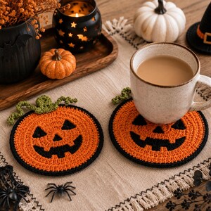 May include: Two round, orange crochet coasters featuring a black jack-o'-lantern face design with a green stem. Each coaster has a black border and is crafted with a textured yarn finish. One coaster supports a ceramic mug filled with a light brown beverage.