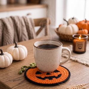 May include: A round, orange and black crocheted coaster featuring a jack-o'-lantern face design with a small green crocheted stem attached to the side, supporting a speckled ceramic mug filled with a steaming dark beverage.