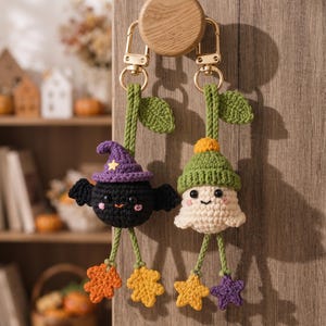 May include: Two crochet keychains hanging from a wooden knob. One is a black bat wearing a purple hat with a yellow star, and the other is a white ghost wearing a green hat. Both have green stems with leaves and dangling colorful leaf-shaped charms.