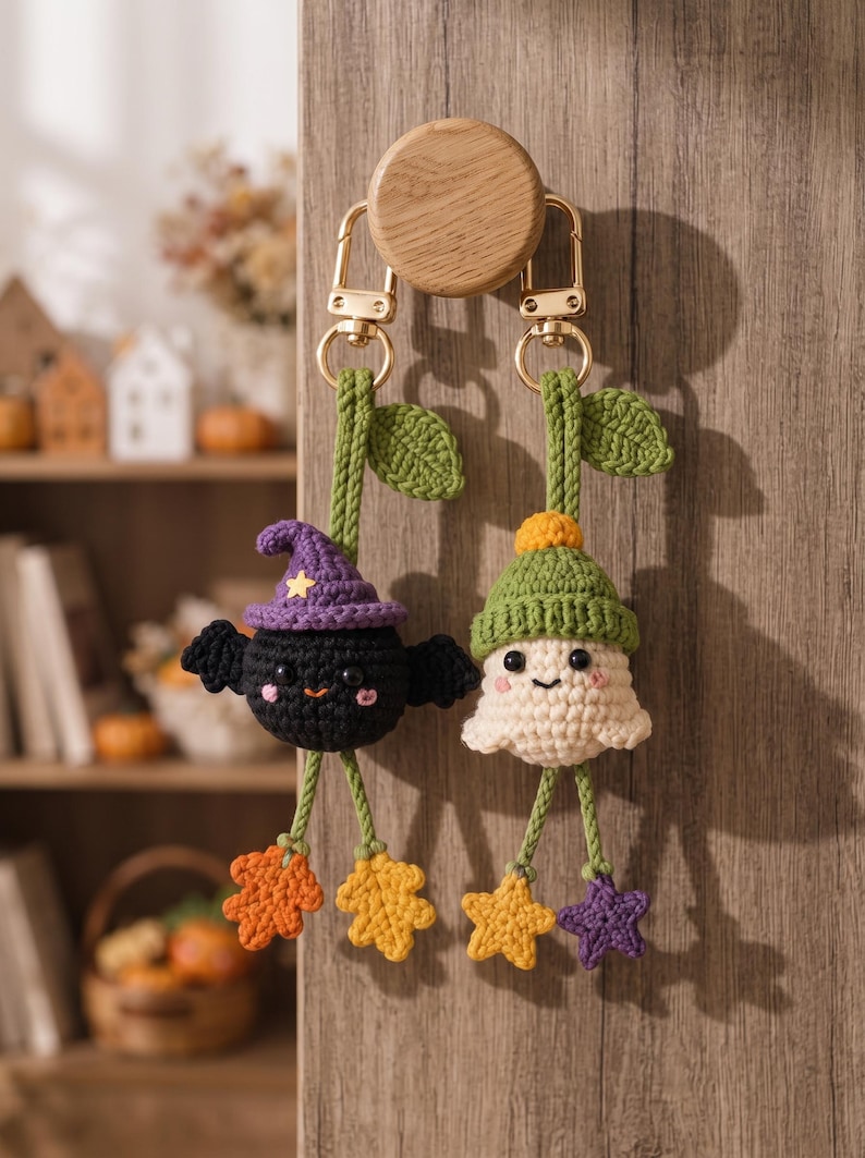 May include: Two crochet keychains hanging from a wooden knob. One is a black bat wearing a purple hat with a yellow star, and the other is a white ghost wearing a green hat. Both have green stems with leaves and dangling colorful leaf-shaped charms.