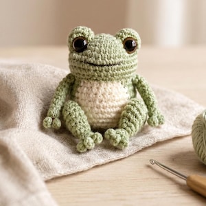 May include: A crocheted frog toy in green and cream tones. The frog has large, dark eyes and is sitting on a beige cloth. A ball of green yarn and a crochet hook are also visible, indicating a craft project.