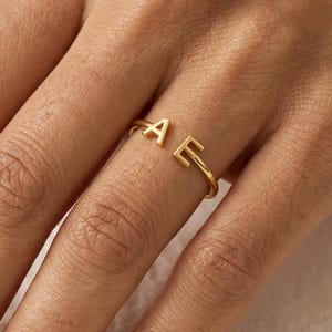 Puede incluir: Un anillo de tono dorado que presenta dos letras mayúsculas, A y E, posicionadas en los extremos abiertos de la banda. El anillo tiene un acabado liso y pulido, con una estructura de metal fina y sencilla que rodea el dedo con una abertura.